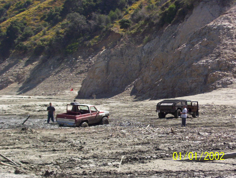 Muddy red pickup truck stuck in mud with a person standing next to it; dark SUV with open doors in background