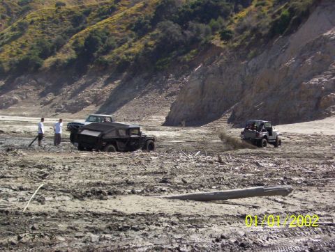 1985 Ford Bronco - Off-road vehicles traverse a challenging muddy landscape with a rocky cliff in the background.