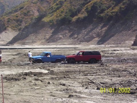1985 Ford Bronco - Two vehicles are stuck in a muddy, dry riverbed with hills in the background.