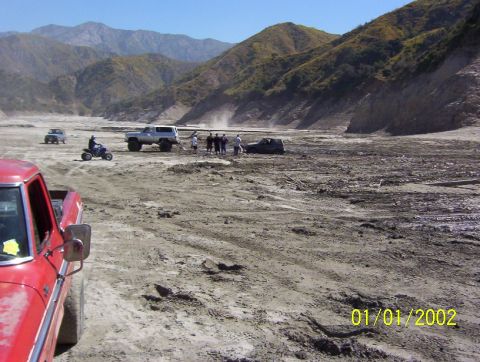 1985 Ford Bronco - A red pickup truck is in the foreground of a scene with multiple vehicles and people in a dry, mountainous landscape.