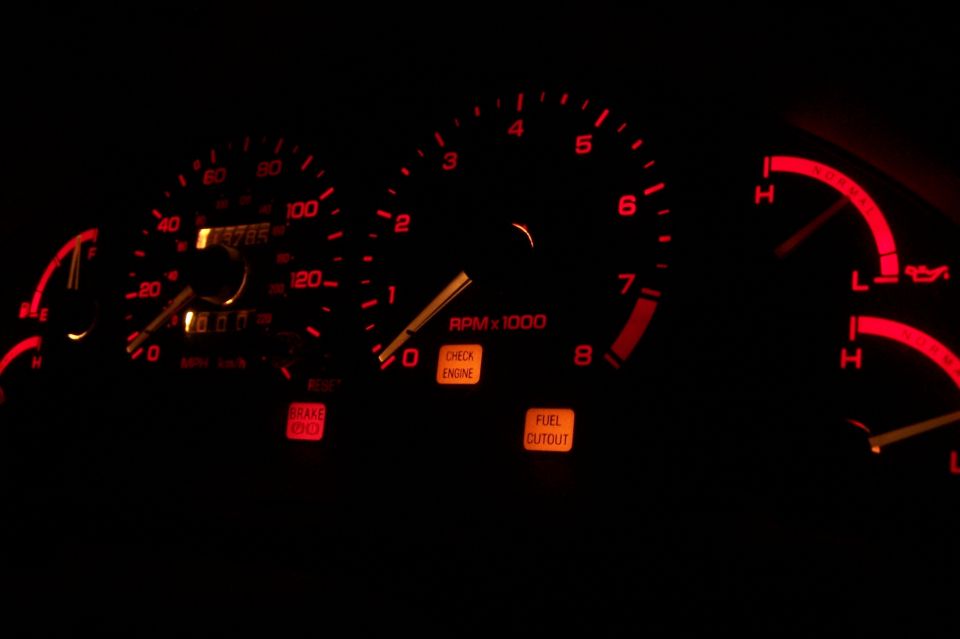 Car dashboard gauges with illuminated red needles and orange warning lights for brake, check engine, and fuel cutout