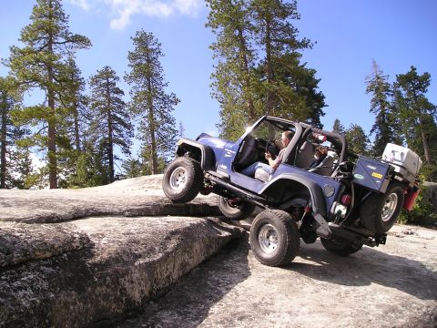 1989 Ford Bronco - A blue Jeep navigates a rocky trail with trees and a clear blue sky in the background.