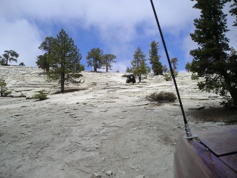 1989 Ford Bronco - An off-road vehicle navigates a steep, rocky terrain with scattered trees and a cloudy blue sky overhead.
