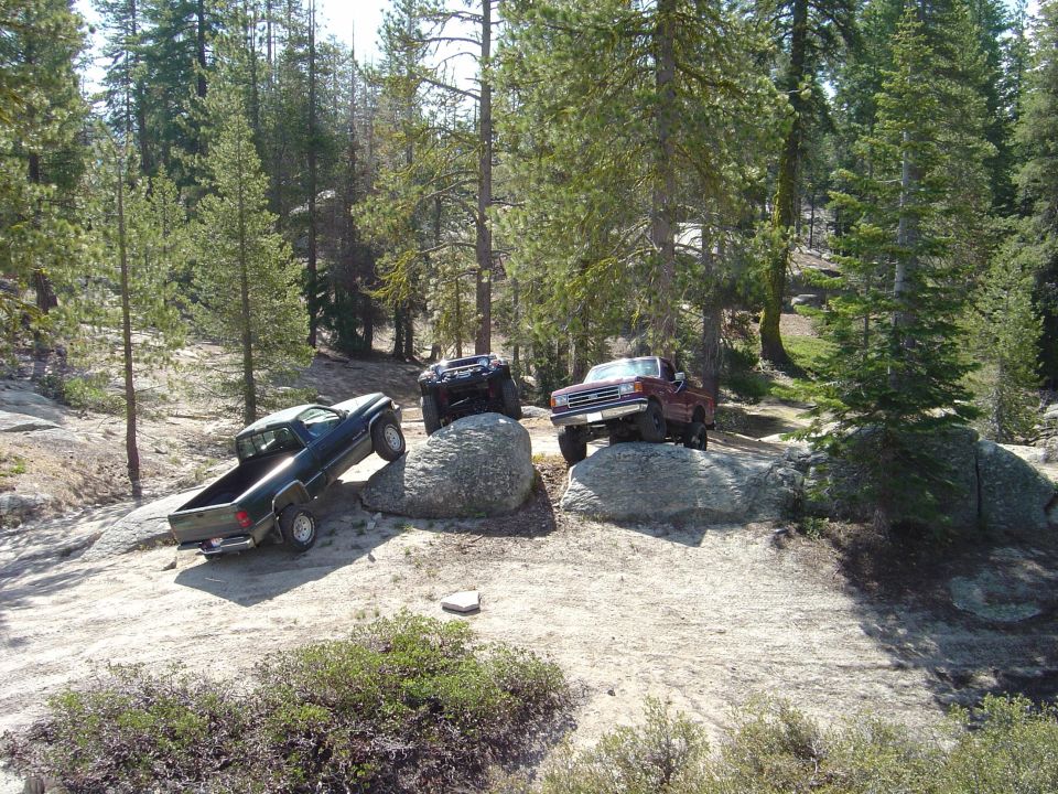 Red pickup truck with lifted suspension on a rock, dark pickup truck on a rock, and black ATV on a rock in a forest