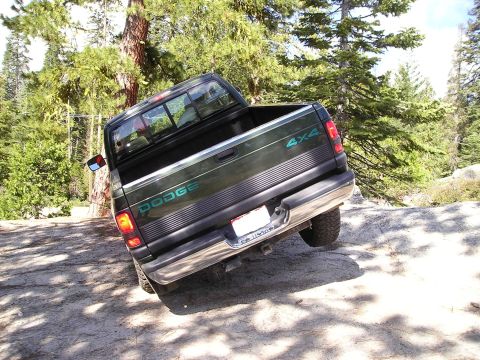 1989 Ford Bronco - Dodge pickup truck navigating an off-road trail with rocky terrain and trees.
