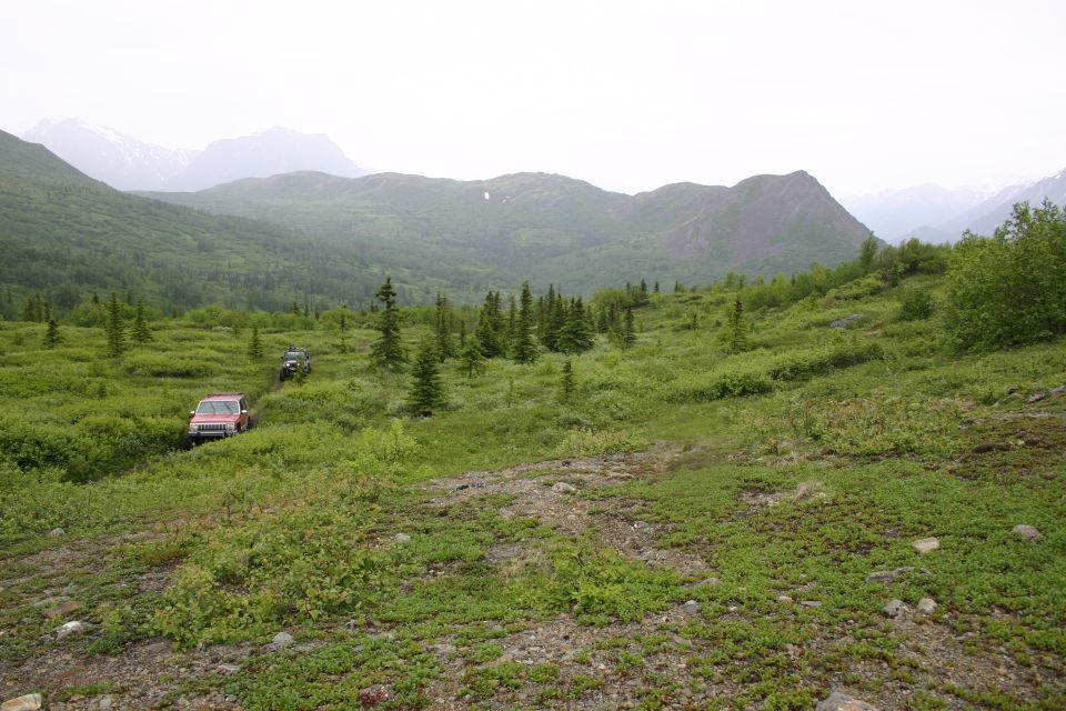 Two off-road vehicles navigate a grassy, hilly terrain with mountains in the background