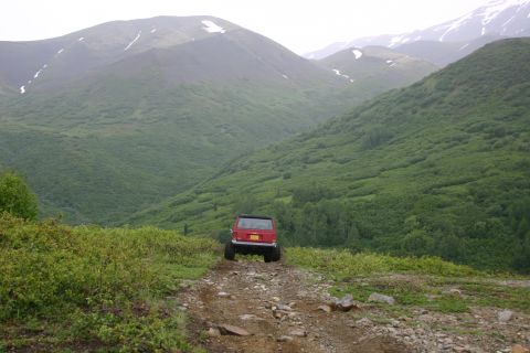 2003 Jeep TJ - A red 2003 Jeep TJ navigates a rocky dirt road through a lush, green mountain valley.
