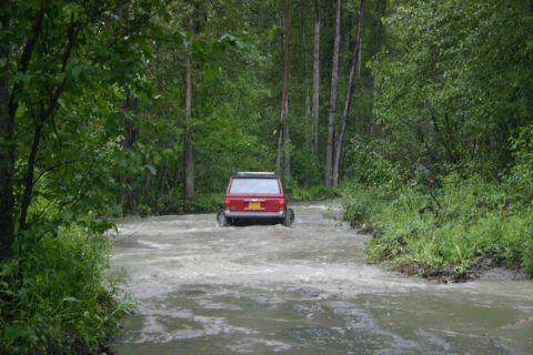 2003 Jeep TJ - A red SUV navigates a water-covered path surrounded by dense green trees.