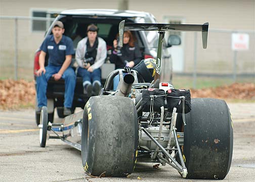 Rear view of a dragster with big slicks and rear wing being towed, people riding on the tow vehicle's tailgate