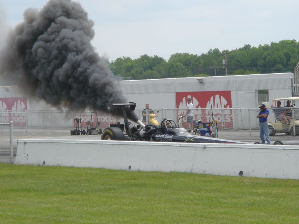 Black Scheid Diesel dragster on a drag strip belching thick black smoke, Mac Tools banners behind