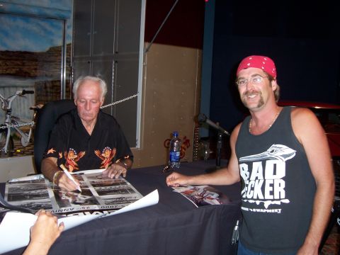 1999 Jeep TJ - Two men interact at a table where one is signing a poster, with a bicycle visible in the background.
