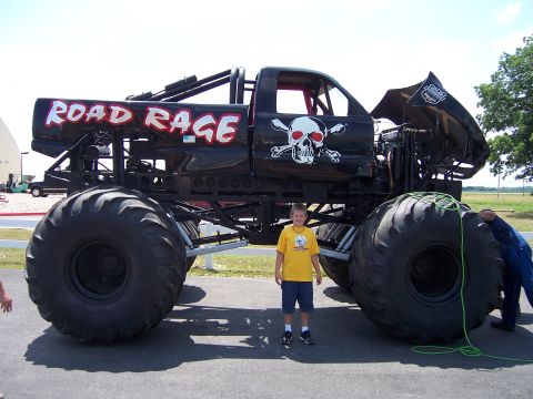 1999 Jeep TJ - A boy stands next to a black monster truck named 'Road Rage', skull and crossbones graphics.