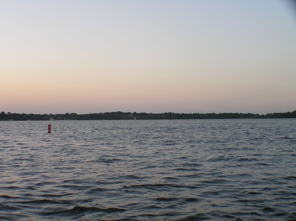 Lake at dusk with red buoy and distant tree-lined shore under a pastel sky
