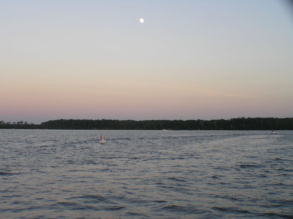 Lake at dusk with moon rising over a tree-lined shore and a buoy in the water