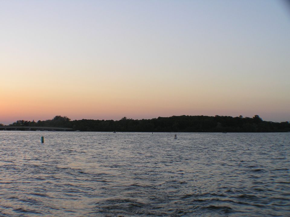 Lake at dusk with tree-lined shore, bridge at left, and orange sunset sky