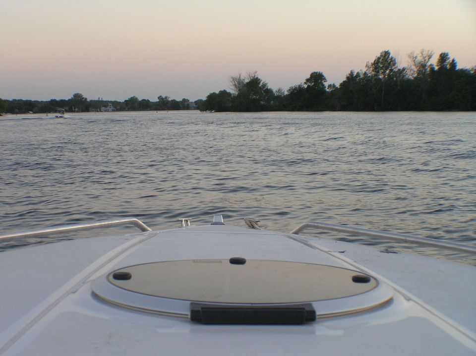 White boat bow deck with hatch, facing calm lake and tree-lined shore at dusk