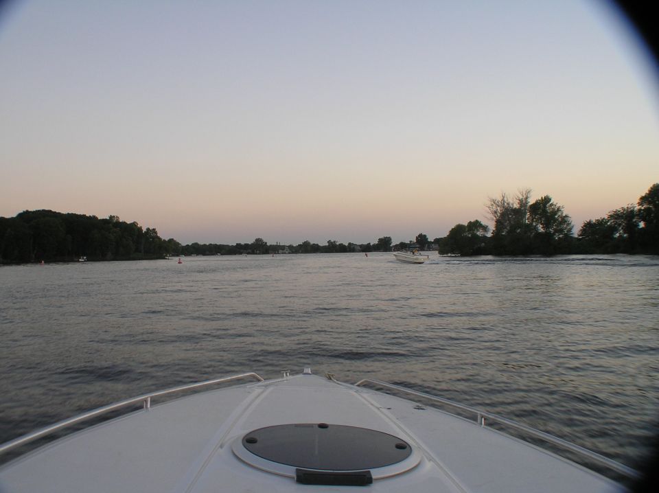 View over white boat bow across calm river at dusk with distant boat and tree-lined shores.