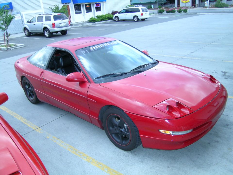Red coupe parked in a lot, with a silver SUV and white van in the background. Text on windshield reads 'Pr...'