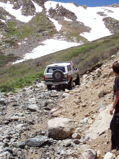 1979 Ford Bronco - A Ford Bronco navigates a challenging, rocky terrain on a mountain trail with snow-capped peaks visible.