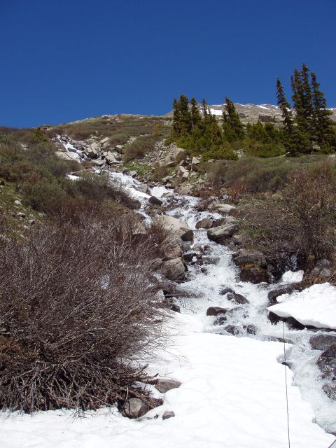 1979 Ford Bronco - A mountain stream cascades over rocks, with patches of snow visible on the banks under a bright blue sky.