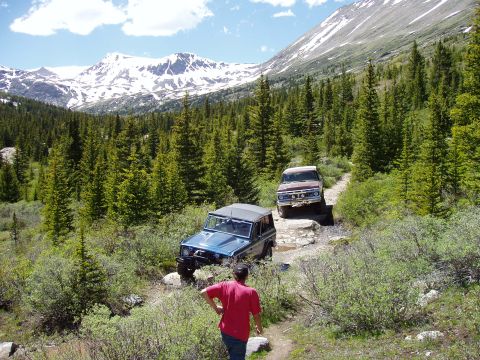1979 Ford Bronco - Two off-road vehicles navigate a challenging mountain trail surrounded by pine trees and snow-capped peaks.