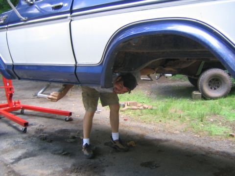 1978 Ford Bronco - A person in shorts bends under the side of a blue-and-white two-tone Ford Bronco propped up in a gravel driveway, working with a length of…