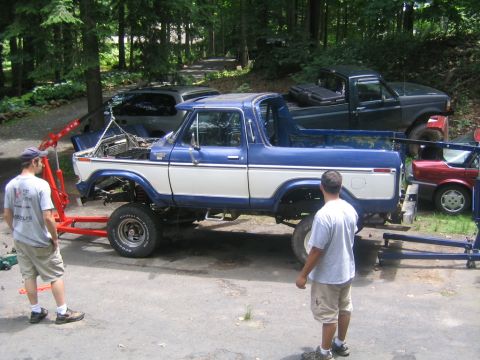 1978 Ford Bronco - Blue and white two-tone Ford Bronco body shell sitting on an engine hoist and stands in a driveway mid-teardown, with the doors, hood, gril…