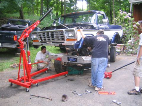 1978 Ford Bronco - A two-tone blue-over-white full-size Ford Bronco up on stands in a wooded driveway with its front bumper, grille and fenders removed for wo…