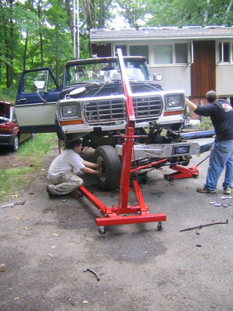 1978 Ford Bronco - Blue and white square-body Ford Bronco hoisted nose-up on a red engine crane in a driveway while two people work on the front axle, engine…