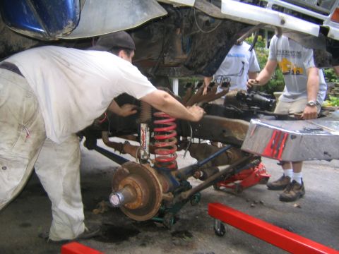 1978 Ford Bronco - Front axle assembly with red coil springs, blue radius arms and a rusty brake rotor being rolled out from under a lifted blue truck body on…