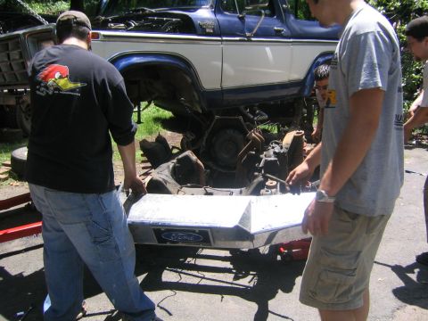 1978 Ford Bronco - Several men lift a bare polished aluminum front bumper with a Ford oval badge away from a blue-and-white full-size Ford Bronco that has bee…