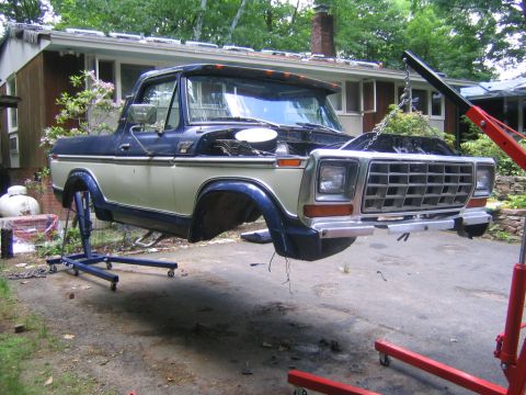 1978 Ford Bronco - A two-tone tan and dark blue full-size Ford Bronco mid-teardown in a driveway, with the removable top and doors off, hood open, and the fro…