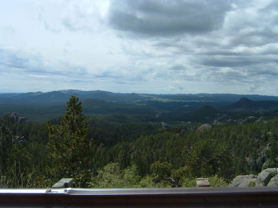 Guardrail in foreground with a vast expanse of forested mountains under a cloudy sky