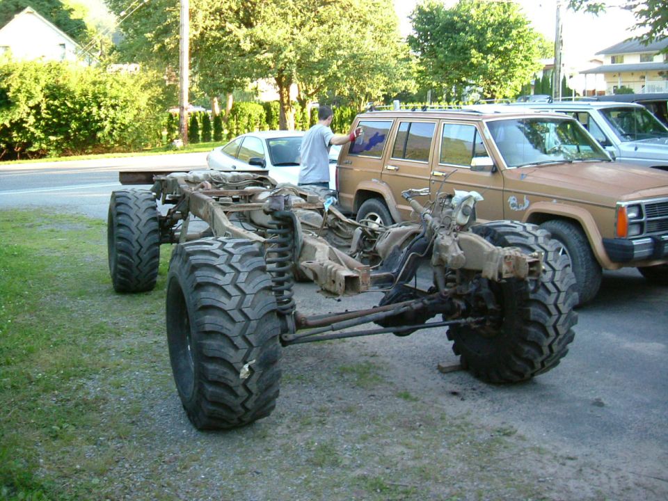 Off-road vehicle chassis with large tires, next to a brown Jeep Cherokee and a white car