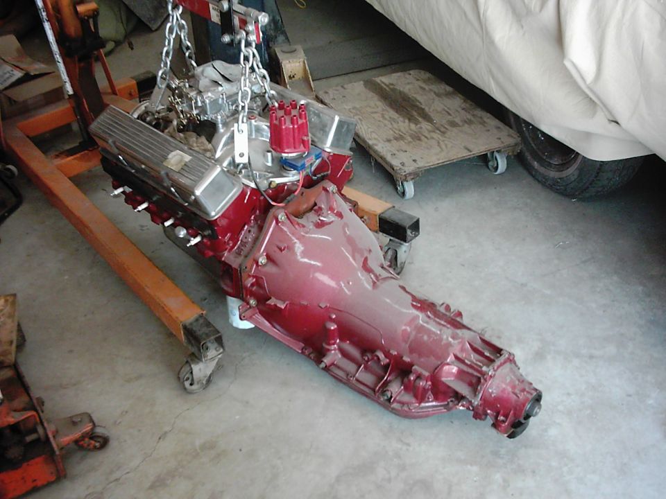 engine and transmission being lifted by an engine hoist. A car covered in a tarp is in the background