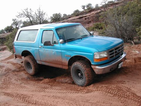 1994 Ford Bronco - A blue Ford Bronco navigates a dusty, off-road trail with red rock formations in the background.