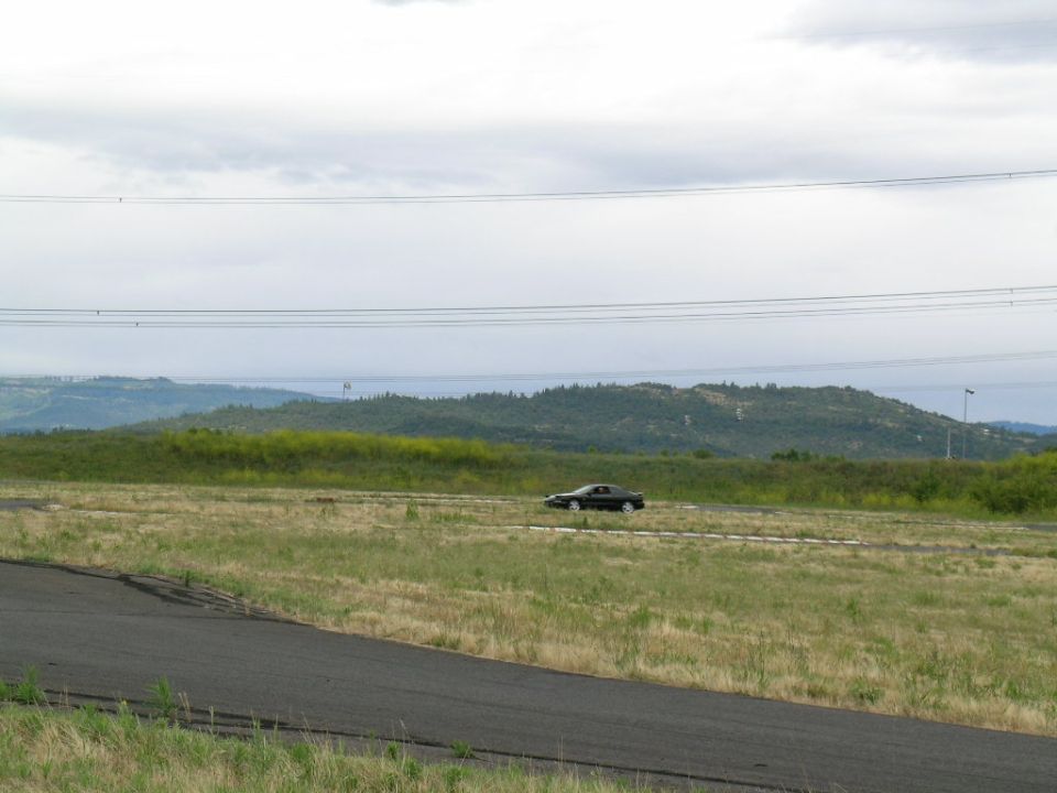 Black 1993 Ford Probe coupe driving on a track with grassy hills and power lines in the background