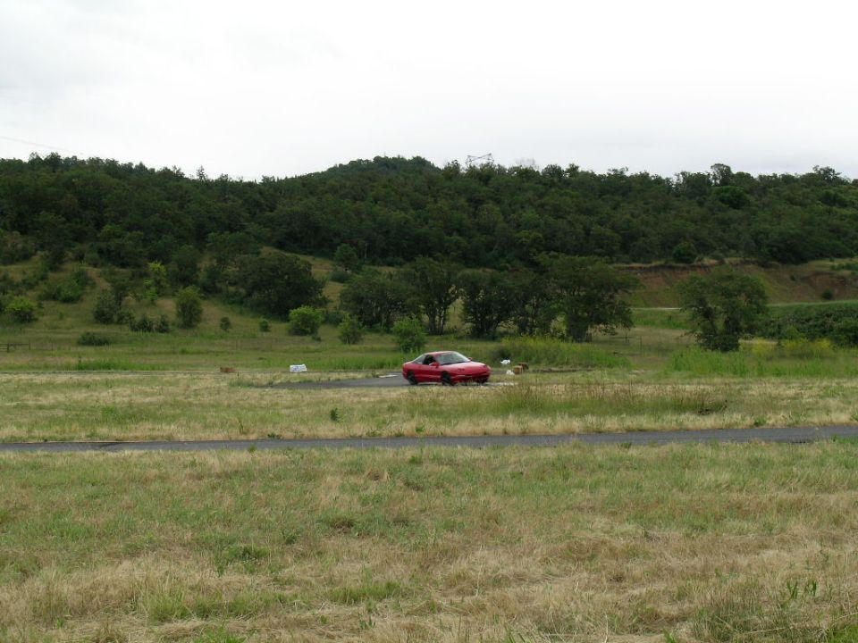 Red coupe on a paved track, 3q-front view. Overcast sky, grassy field, and wooded hill in background