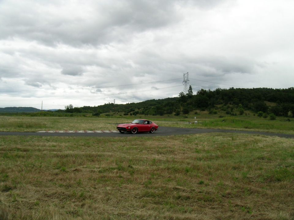 Red Datsun 280Z on a paved track with grassy fields and hills in the background