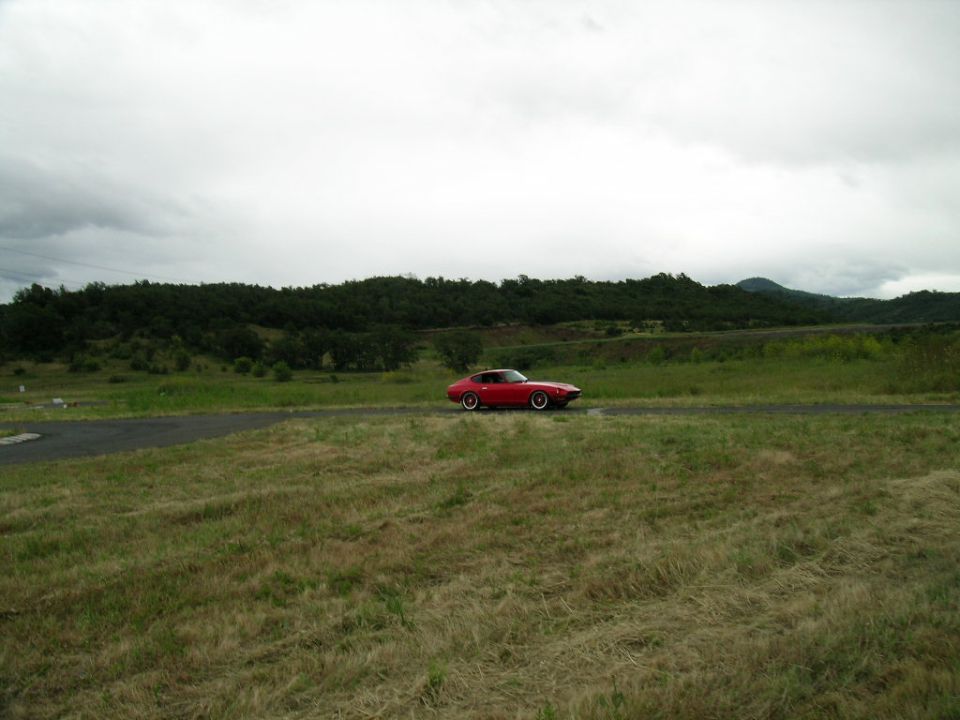 red sports car driving on a road with grassy fields and trees under a cloudy sky