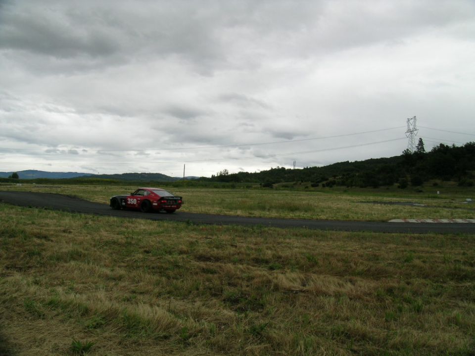 Red muscle car with number 350 on its side drives on a paved track through a grassy field under a cloudy sky