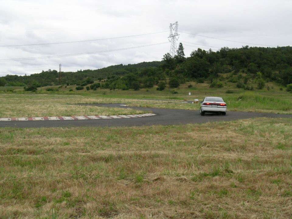 White sedan driving on a race track with grassy hills and a power line tower in the background