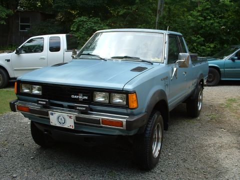 1985 Ford Bronco - Light blue Datsun 4x4 regular-cab pickup shot in 3/4 front view on a gravel driveway, showing the black grille with DATSUN lettering, quad…