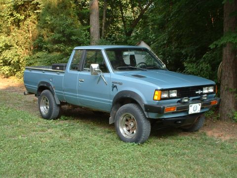 1985 Ford Bronco - A light blue Datsun extended-cab pickup truck photographed from the front-right 3/4 angle, parked on grass near a wooded area. It has a lif…