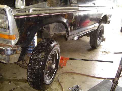 1979 Ford Bronco - A black late-1970s Ford Bronco is jacked up on a garage floor with the driver's front wheel pulled off and leaned against the body, exposin…