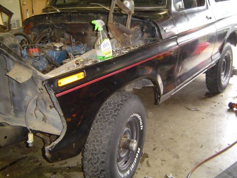 1979 Ford Bronco - Black late-1970s Ford Bronco in a shop with the hood, grille and front clip disassembled, exposing a blue V8 engine, valve covers and the r…