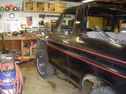 1979 Ford Bronco - Driver-side profile of a black late-1970s Ford Bronco inside a home shop with the doors, front fender and hood removed and lengths of trim…