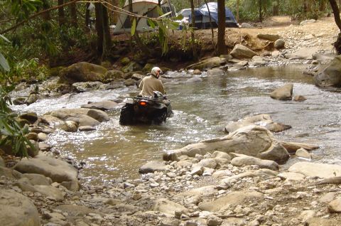 1993 Ford Bronco - An ATV navigates a shallow, rocky river in a wooded area with tents visible in the background.