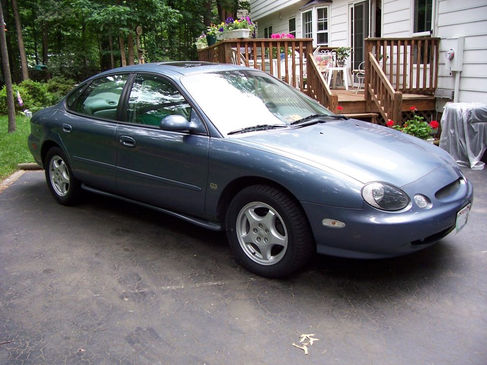 Blue sedan parked in driveway, front quarter view. House and trees in background