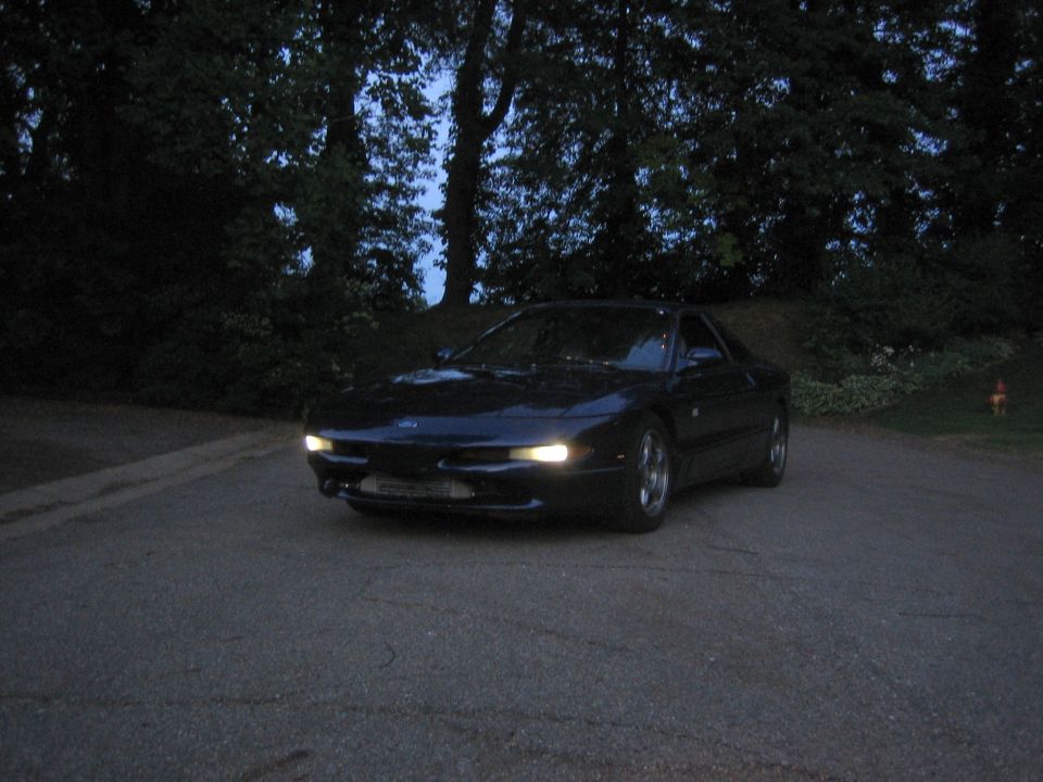 Dark Ford Probe coupe at dusk with headlights on, parked on asphalt by woods
